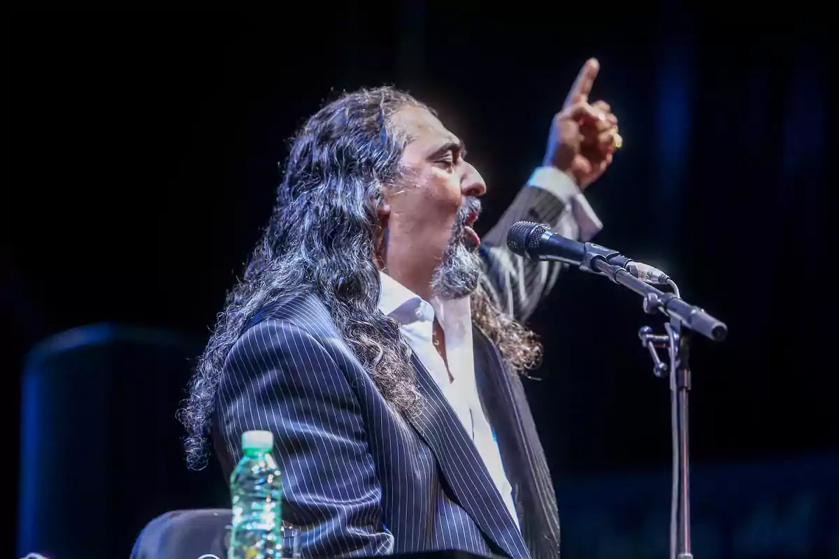 Hombre con cabello largo y barba cantando apasionadamente frente a un micrófono mientras levanta una mano señalando hacia arriba Hombre con cabello largo y barba cantando apasionadamente frente a un micrófono mientras levanta una mano señalando hacia arriba