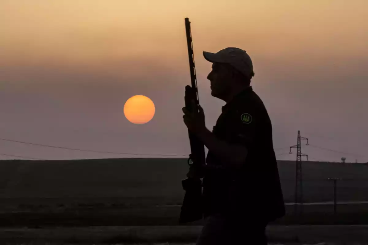 Hombre con gorra sosteniendo un rifle mientras camina al atardecer en un paisaje rural Hombre con gorra sosteniendo un rifle mientras camina al atardecer en un paisaje rural
