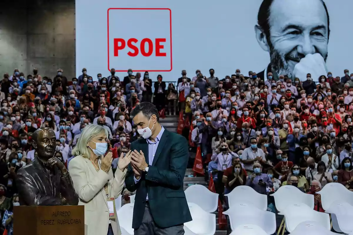 Un auditorio lleno de personas con mascarilla aplaude mientras dos personas conversan junto a un busto y un gran cartel de PSOE, al fondo se ve una imagen en blanco y negro de un hombre sonriente. Un auditorio lleno de personas con mascarilla aplaude mientras dos personas conversan junto a un busto y un gran cartel de PSOE, al fondo se ve una imagen en blanco y negro de un hombre sonriente.