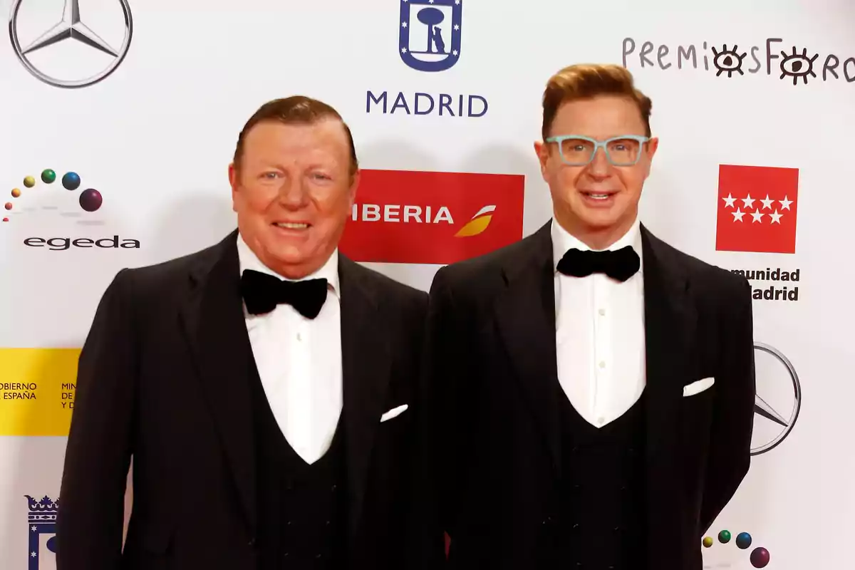 Dos hombres vestidos de esmoquin posan sonrientes en una alfombra roja frente a un fondo con logotipos de patrocinadores y premios. Dos hombres vestidos de esmoquin posan sonrientes en una alfombra roja frente a un fondo con logotipos de patrocinadores y premios.