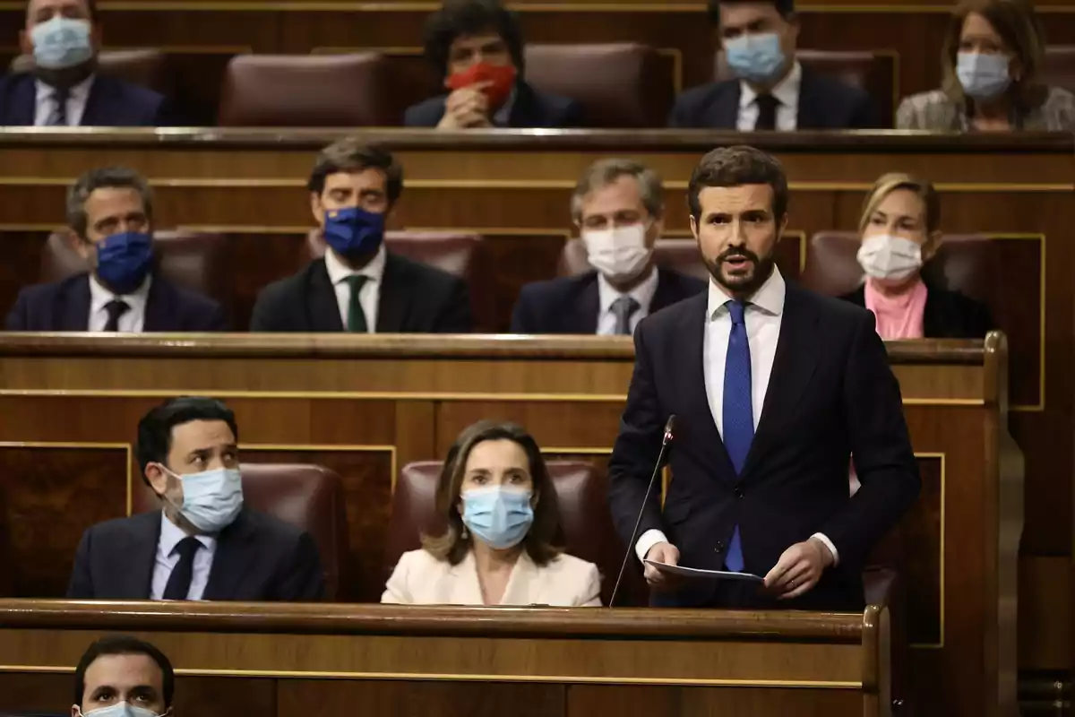 Un hombre de traje y corbata azul habla de pie en un parlamento mientras varias personas con mascarilla lo observan desde sus asientos Un hombre de traje y corbata azul habla de pie en un parlamento mientras varias personas con mascarilla lo observan desde sus asientos