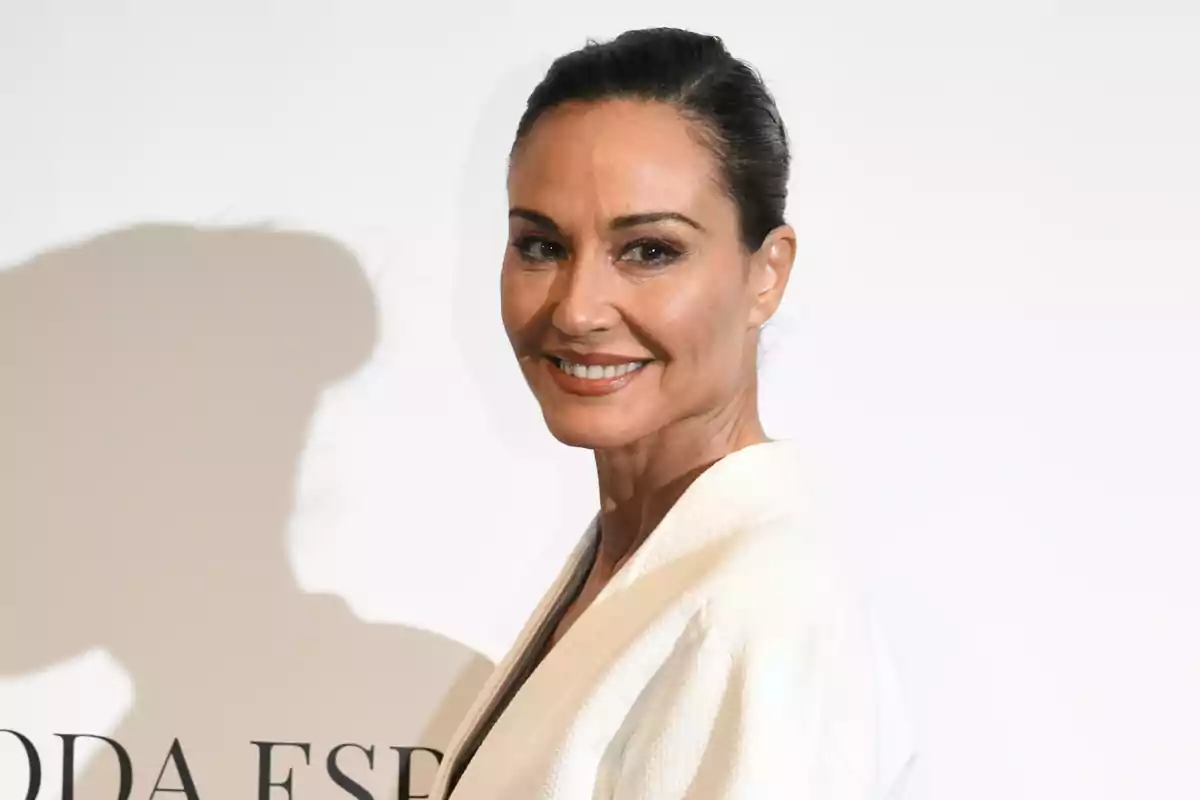 Mujer de cabello oscuro recogido y sonrisa, vestida con ropa blanca, posando frente a un fondo claro Mujer de cabello oscuro recogido y sonrisa, vestida con ropa blanca, posando frente a un fondo claro