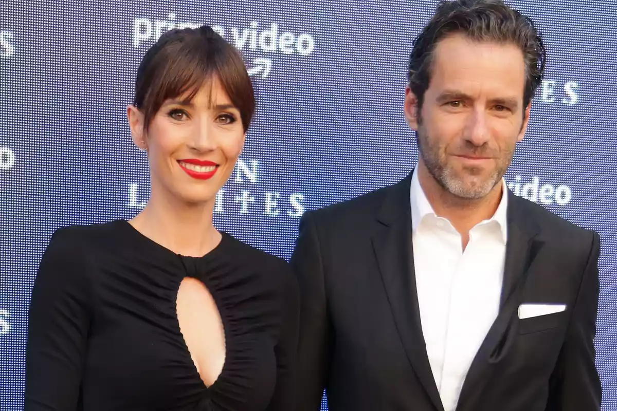 Una mujer con vestido negro y un hombre con traje posan juntos en un evento frente a un fondo con el logo de Prime Video.
