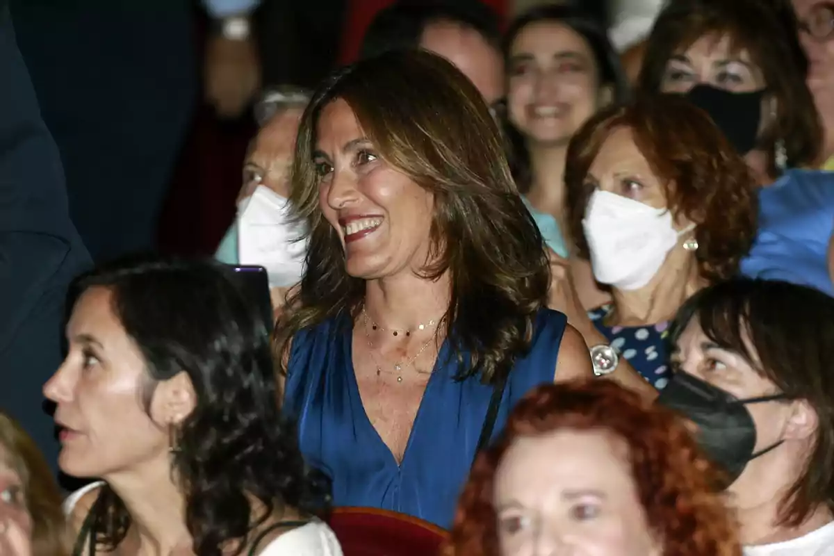 Mujer sonriente con vestido azul sentada entre otras personas en un evento algunas de las cuales llevan mascarilla Mujer sonriente con vestido azul sentada entre otras personas en un evento algunas de las cuales llevan mascarilla