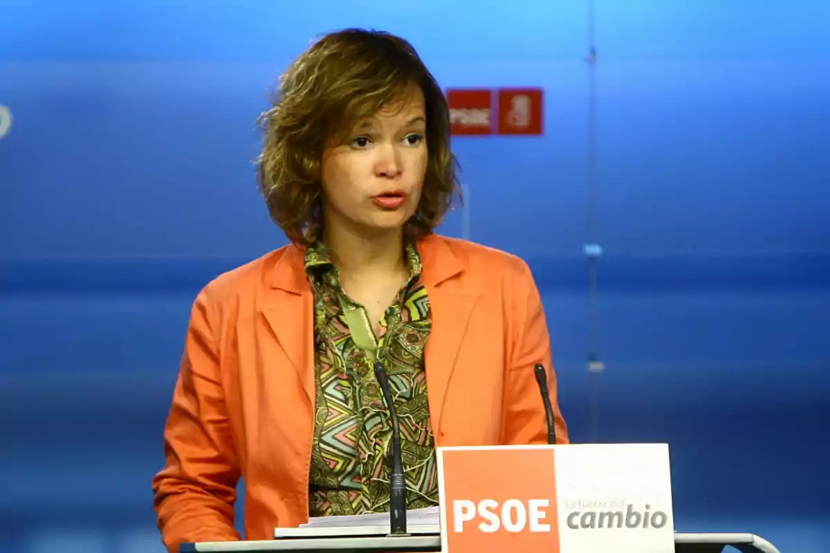Mujer con chaqueta naranja hablando en un atril con el logo del PSOE y fondo azul