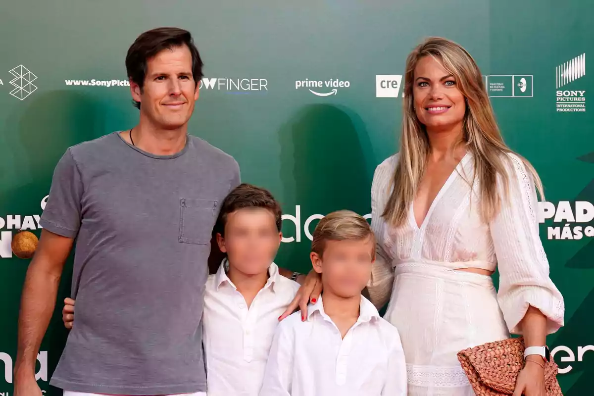 A family of four smiles in front of a green background at an event, with the adults on the sides and the children in the center.