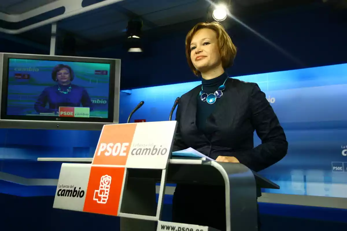 Mujer de pie en un atril con logotipos del PSOE en una sala de prensa con fondo azul y una pantalla mostrando la misma escena Mujer de pie en un atril con logotipos del PSOE en una sala de prensa con fondo azul y una pantalla mostrando la misma escena
