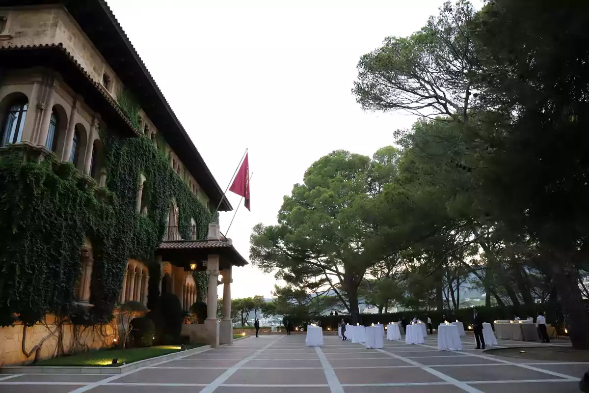 Edificio cubierto de hiedra con una bandera roja en la entrada y varias mesas altas con manteles blancos dispuestas en un patio al aire libre rodeado de árboles Edificio cubierto de hiedra con una bandera roja en la entrada y varias mesas altas con manteles blancos dispuestas en un patio al aire libre rodeado de árboles