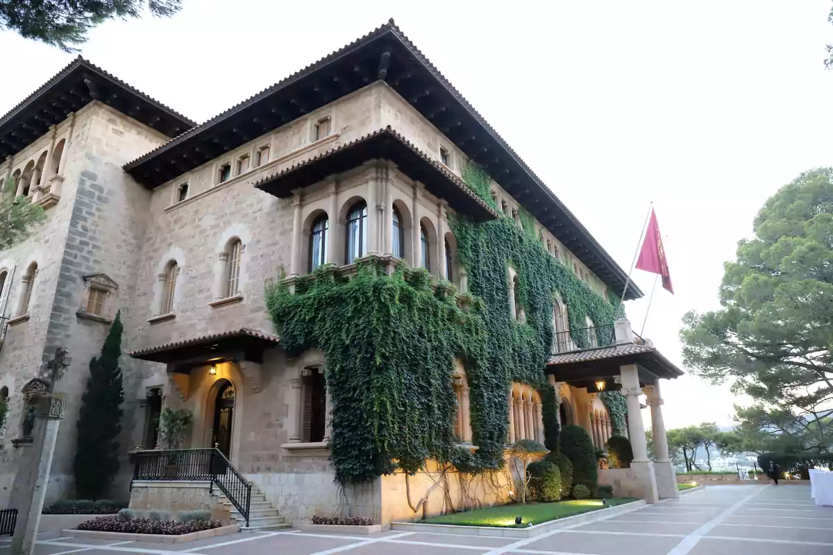 Edificio antiguo de piedra con fachada cubierta de enredaderas verdes y una bandera roja ondeando en la entrada rodeado de árboles y jardines bien cuidados Edificio antiguo de piedra con fachada cubierta de enredaderas verdes y una bandera roja ondeando en la entrada rodeado de árboles y jardines bien cuidados