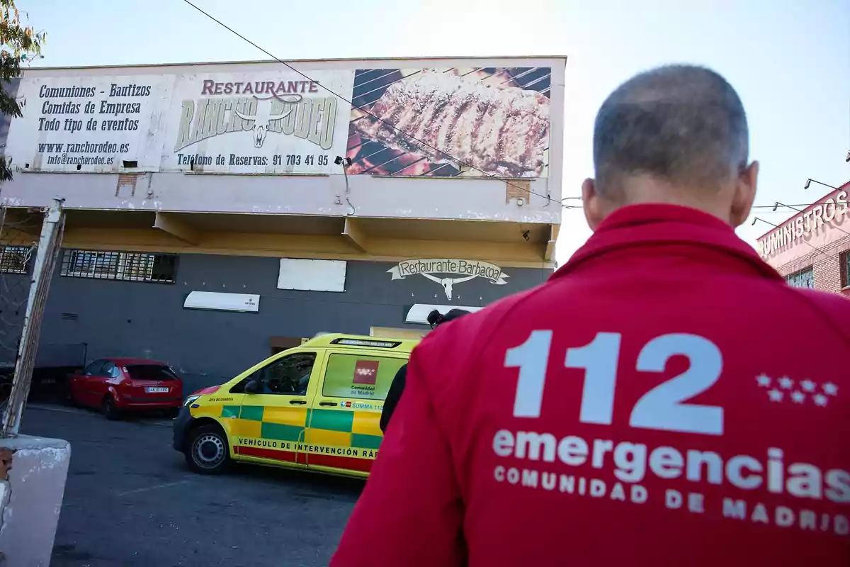 Un trabajador de emergencias con chaqueta roja observa una ambulancia amarilla estacionada frente a un restaurante de barbacoa llamado Rancho Rodeo Un trabajador de emergencias con chaqueta roja observa una ambulancia amarilla estacionada frente a un restaurante de barbacoa llamado Rancho Rodeo