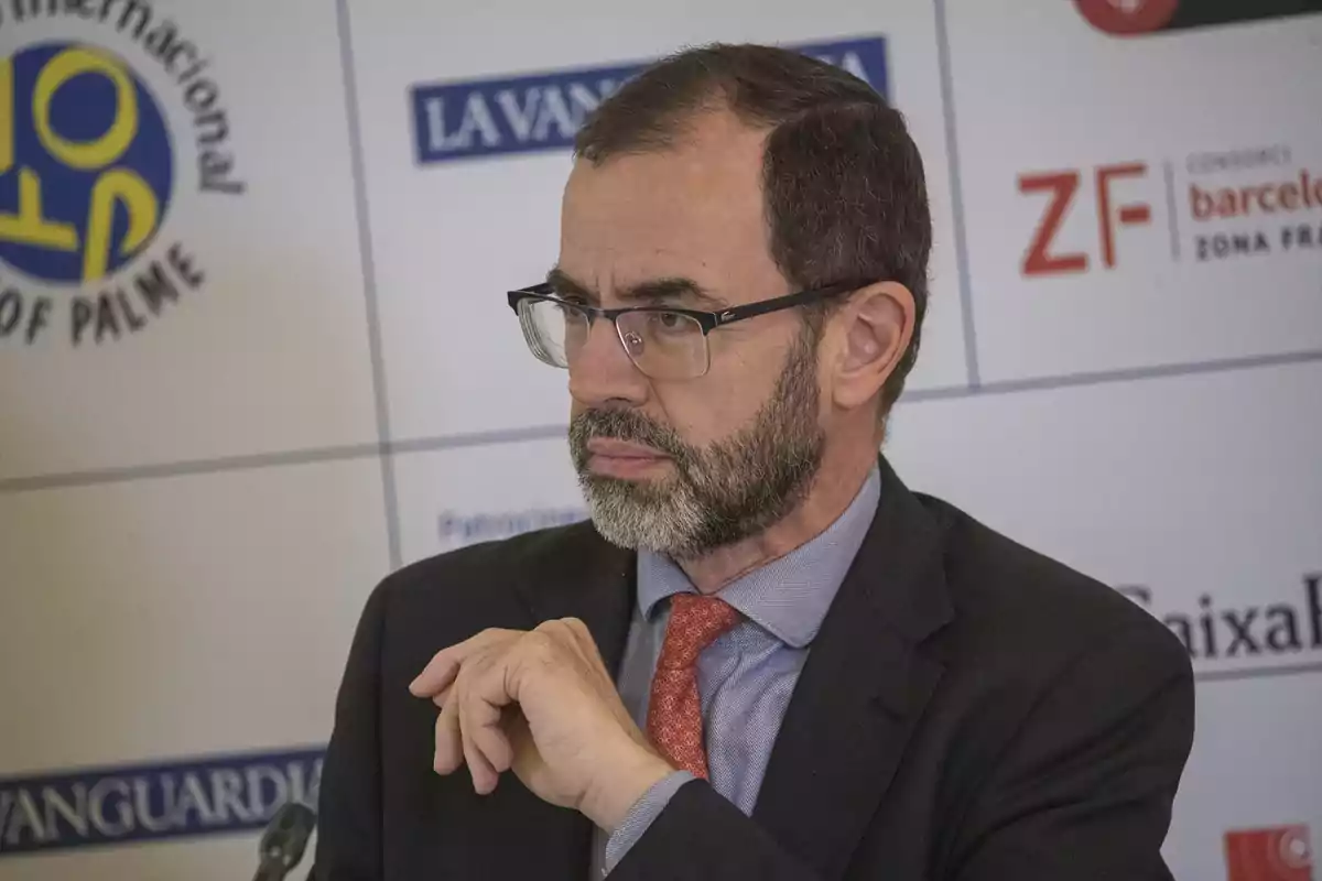 Hombre de mediana edad con barba y gafas, vestido con traje y corbata roja, sentado frente a un fondo con logotipos de empresas y medios de comunicación Hombre de mediana edad con barba y gafas, vestido con traje y corbata roja, sentado frente a un fondo con logotipos de empresas y medios de comunicación
