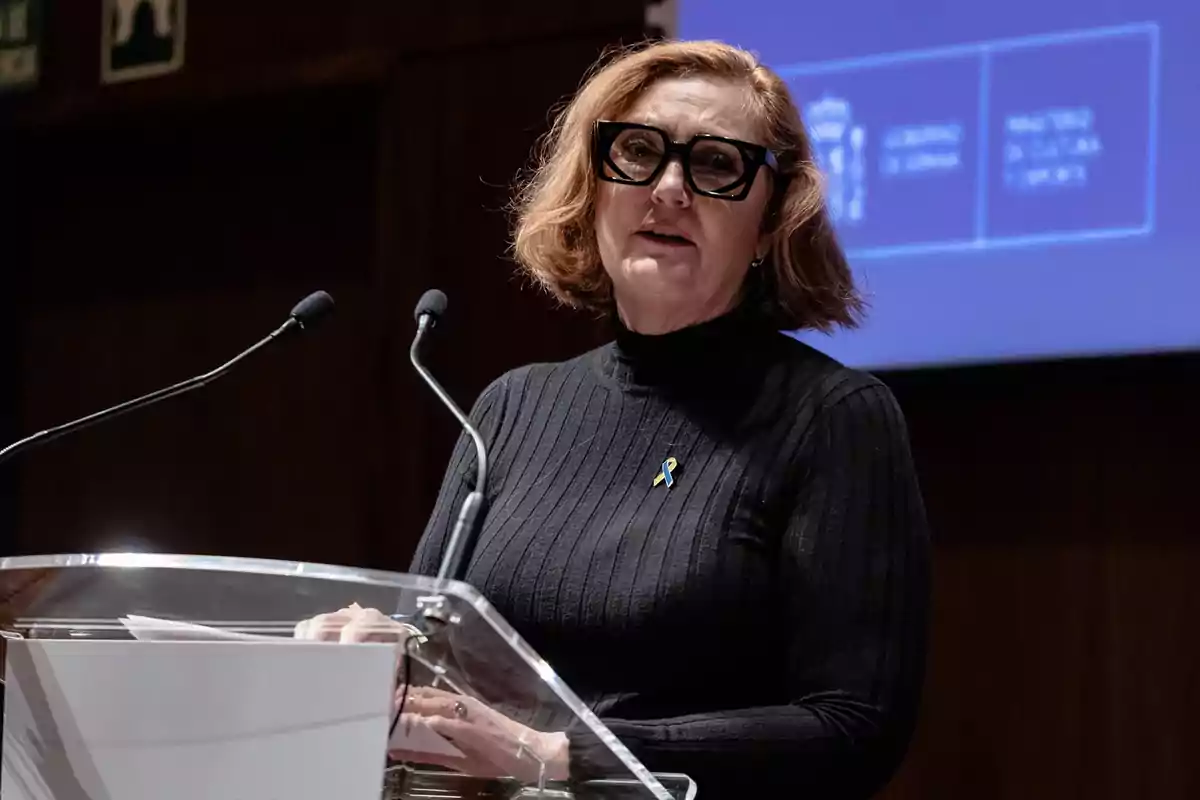Mujer de cabello corto y gafas grandes hablando en un podio con dos micrófonos y fondo azul con logotipos institucionales Mujer de cabello corto y gafas grandes hablando en un podio con dos micrófonos y fondo azul con logotipos institucionales