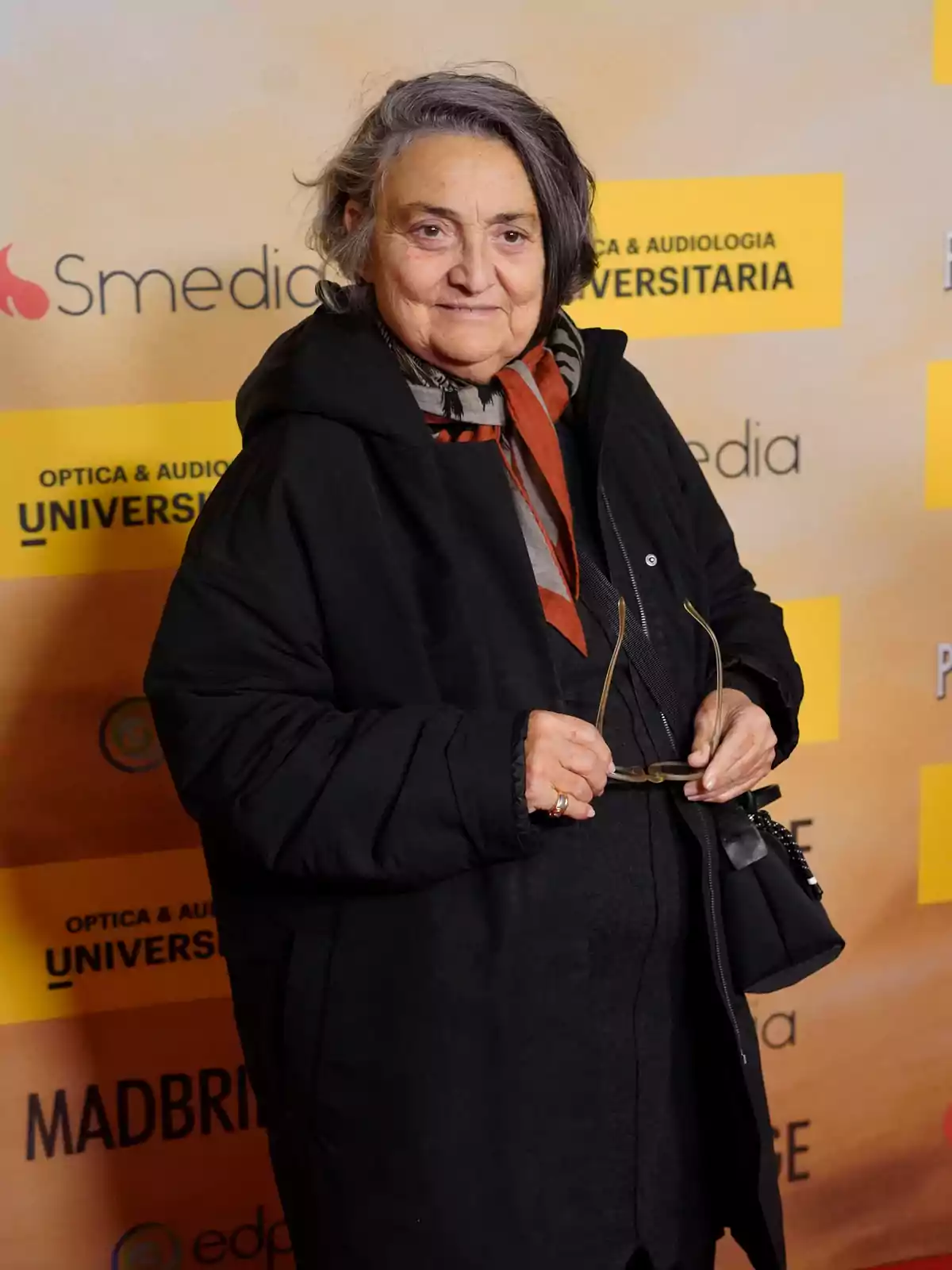 Mujer mayor de cabello canoso y corto vestida con abrigo negro y bufanda, sosteniendo unas gafas en la mano, posando frente a un fondo con logotipos y texto amarillo Mujer mayor de cabello canoso y corto vestida con abrigo negro y bufanda, sosteniendo unas gafas en la mano, posando frente a un fondo con logotipos y texto amarillo