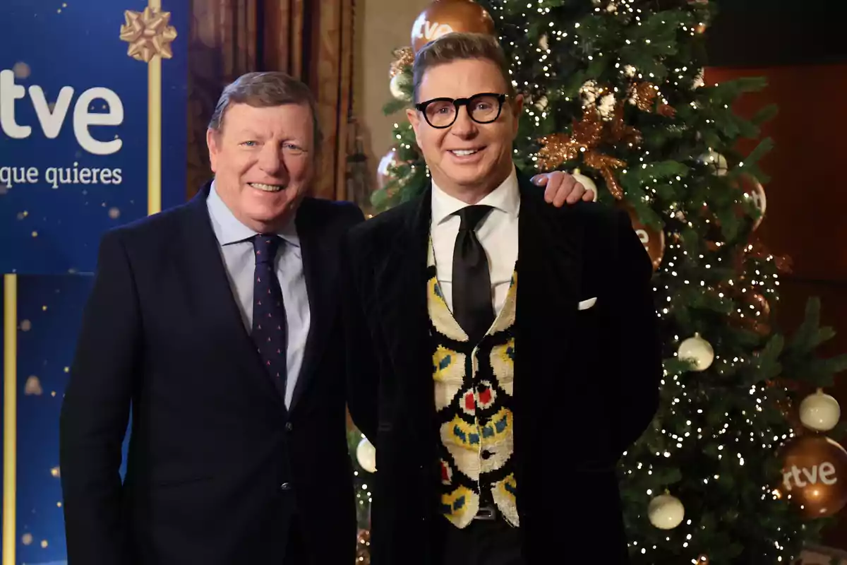 Dos hombres vestidos de traje posan sonrientes frente a un árbol de Navidad decorado y un cartel de RTVE Dos hombres vestidos de traje posan sonrientes frente a un árbol de Navidad decorado y un cartel de RTVE