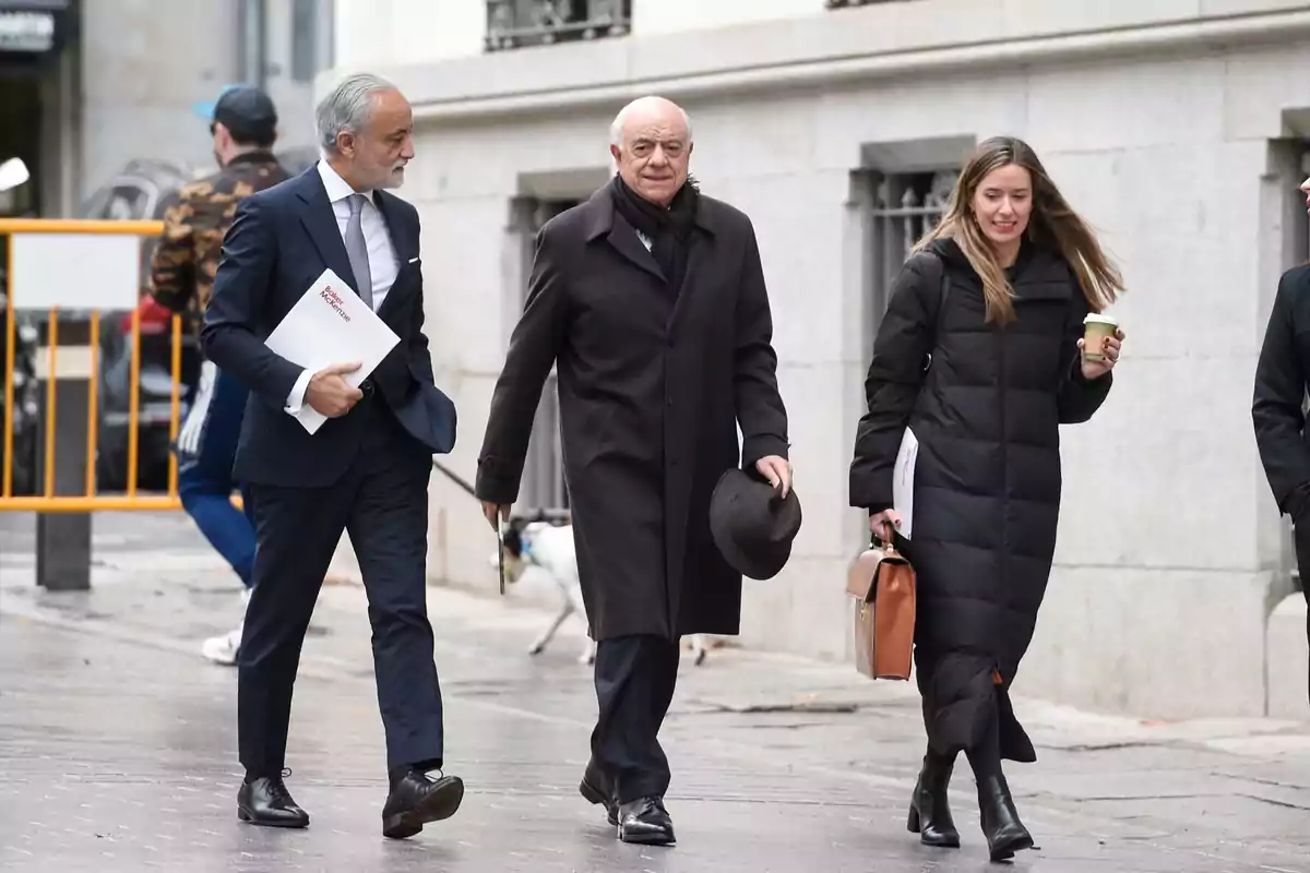 Tres personas elegantemente vestidas caminan por la acera en un día frío, una de ellas lleva un café y otra un sombrero en la mano Tres personas elegantemente vestidas caminan por la acera en un día frío, una de ellas lleva un café y otra un sombrero en la mano