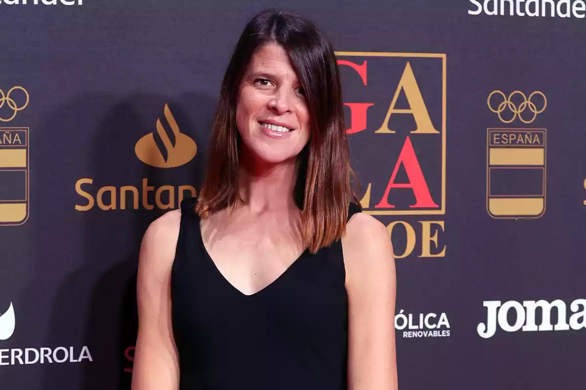 Mujer con vestido negro posando sonriente en una alfombra roja con fondo de logotipos y letras doradas y rojas