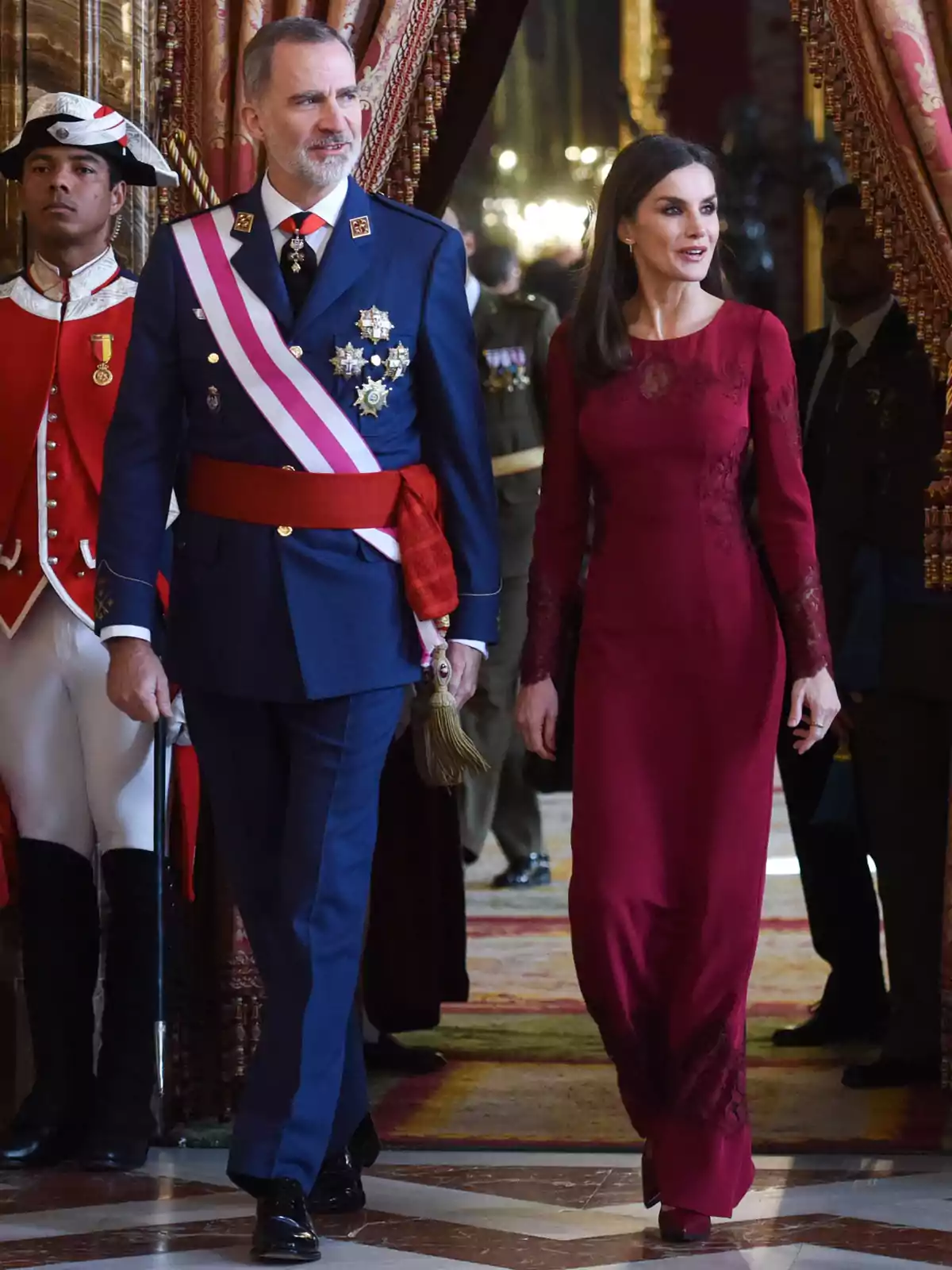 Un hombre con uniforme azul condecorado y una mujer con vestido largo burdeos caminan juntos en un evento formal rodeados de guardias y cortinas elegantes Un hombre con uniforme azul condecorado y una mujer con vestido largo burdeos caminan juntos en un evento formal rodeados de guardias y cortinas elegantes