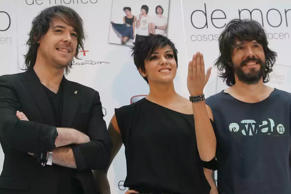 Tres personas posan sonrientes frente a un fondo promocional, dos hombres a los lados y una mujer en el centro con el brazo levantado Tres personas posan sonrientes frente a un fondo promocional, dos hombres a los lados y una mujer en el centro con el brazo levantado