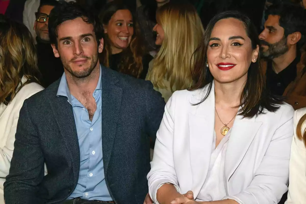 Un hombre y una mujer sentados juntos en un evento social, ambos vestidos de manera elegante y rodeados de otras personas que conversan y sonríen. Un hombre y una mujer sentados juntos en un evento social, ambos vestidos de manera elegante y rodeados de otras personas que conversan y sonríen.