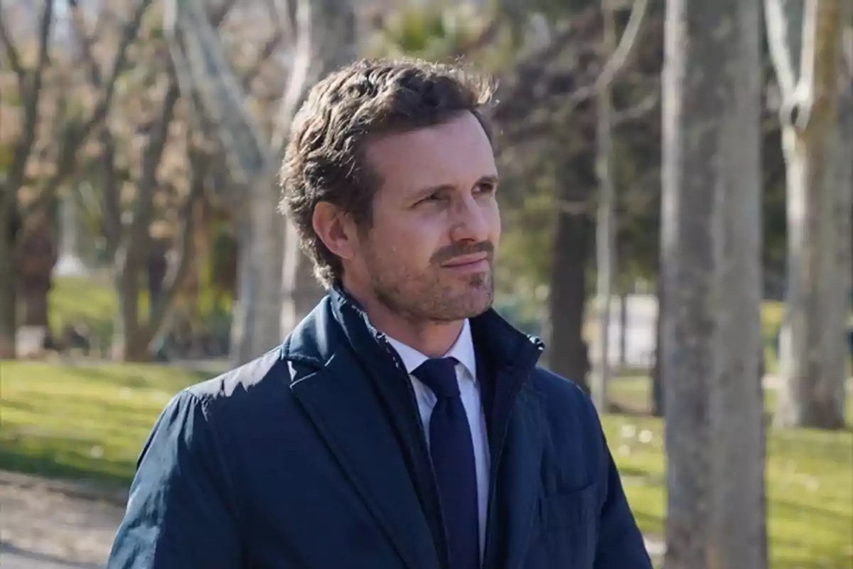 Hombre de cabello corto y barba ligera vistiendo abrigo azul y corbata en un parque con árboles al fondo Hombre de cabello corto y barba ligera vistiendo abrigo azul y corbata en un parque con árboles al fondo
