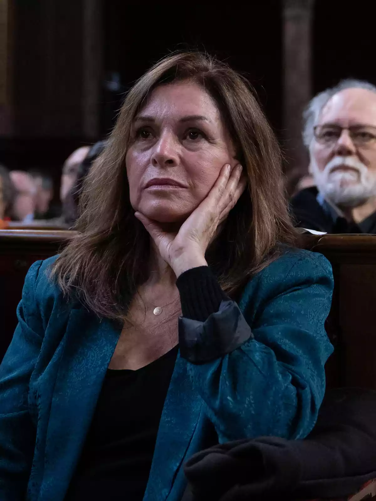 Mujer de cabello castaño sentada con la mano apoyada en la mejilla, vestida con un saco azul, observando atentamente algo frente a ella en un ambiente interior con otras personas al fondo. Mujer de cabello castaño sentada con la mano apoyada en la mejilla, vestida con un saco azul, observando atentamente algo frente a ella en un ambiente interior con otras personas al fondo.