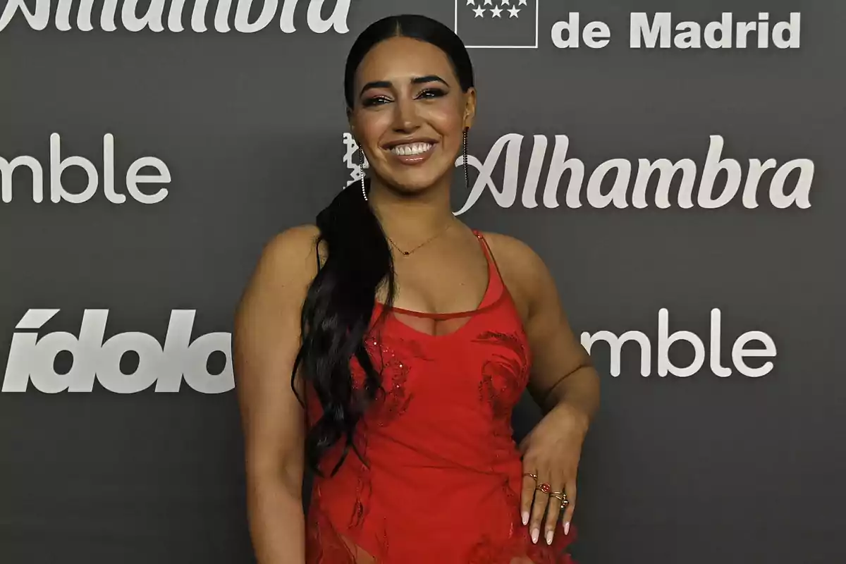 Mujer sonriendo con vestido rojo posando en una alfombra roja frente a un fondo con logotipos blancos
