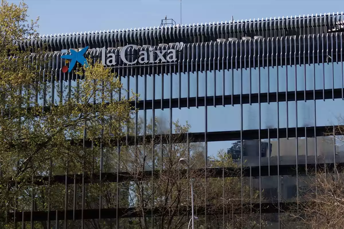Edificio moderno de cristal con el logo de la Caixa en la parte superior y árboles en primer plano Edificio moderno de cristal con el logo de la Caixa en la parte superior y árboles en primer plano