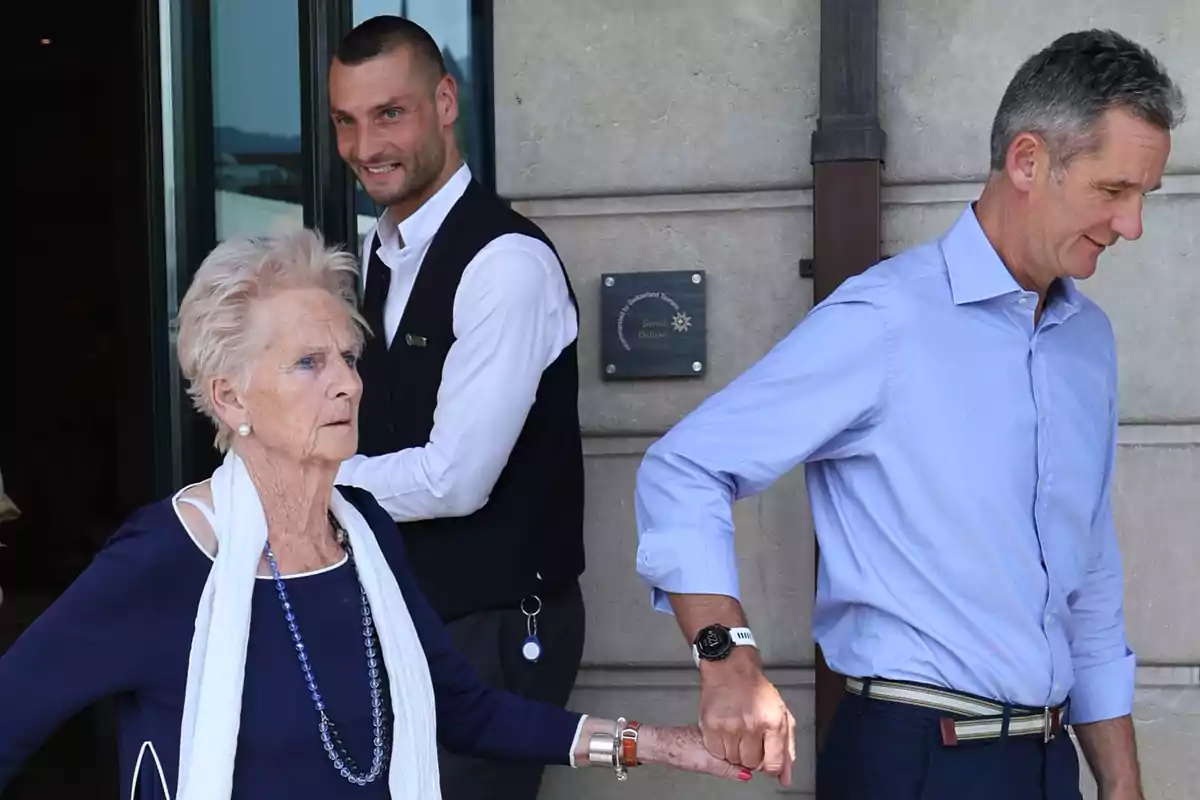 Un hombre de camisa azul ayuda a una mujer mayor vestida de azul y blanco a caminar mientras un hombre con chaleco oscuro los observa y sonríe en la entrada de un edificio. Un hombre de camisa azul ayuda a una mujer mayor vestida de azul y blanco a caminar mientras un hombre con chaleco oscuro los observa y sonríe en la entrada de un edificio.