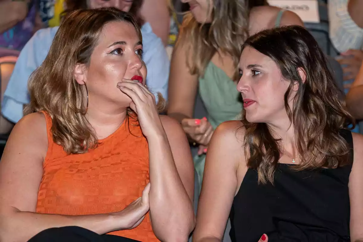 Dos mujeres sentadas conversan en un evento, una lleva blusa naranja y la otra vestido negro, ambas parecen concentradas en la charla Dos mujeres sentadas conversan en un evento, una lleva blusa naranja y la otra vestido negro, ambas parecen concentradas en la charla