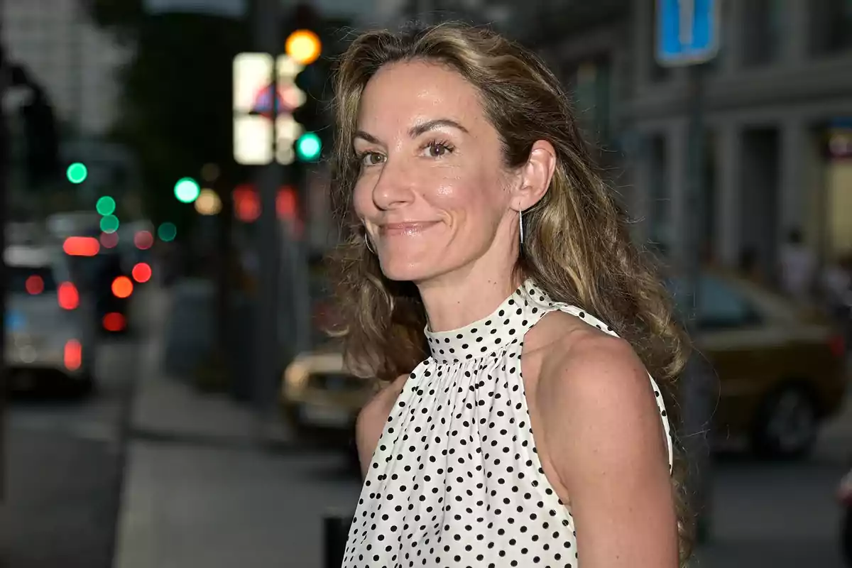Long-haired, wavy-haired woman wearing a white blouse with black polka dots, smiling on a city street at sunset
