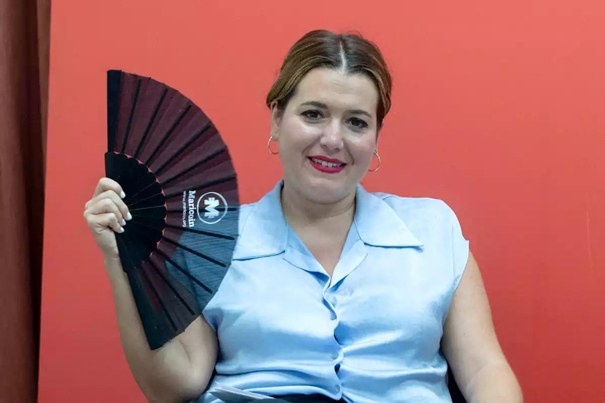 Mujer sonriente con blusa celeste sostiene un abanico negro frente a un fondo rojo Mujer sonriente con blusa celeste sostiene un abanico negro frente a un fondo rojo