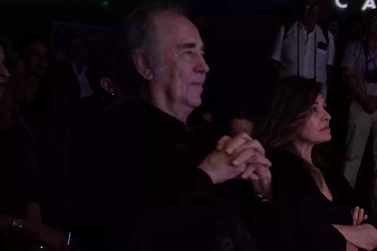 Dos personas sentadas en un lugar oscuro observan atentamente hacia el frente rodeadas de otras personas de pie al fondo Dos personas sentadas en un lugar oscuro observan atentamente hacia el frente rodeadas de otras personas de pie al fondo