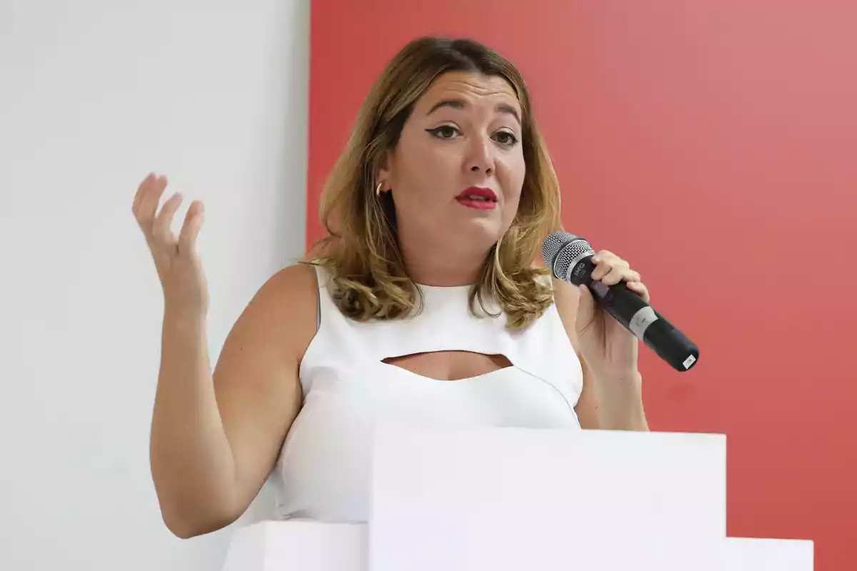 Mujer de cabello rubio hablando por micrófono y gesticulando con la mano frente a un fondo rojo y blanco