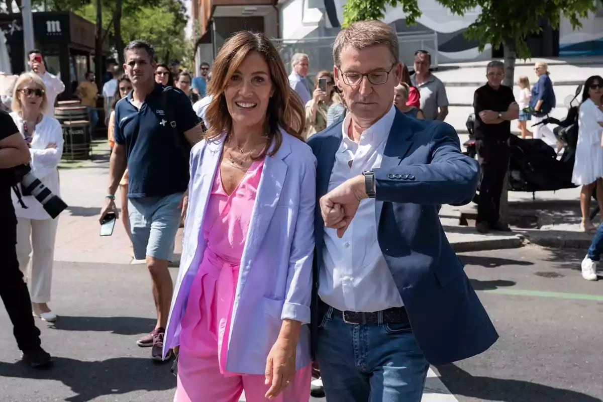 Una pareja camina por la calle rodeada de gente mientras el hombre mira la hora en su reloj y la mujer sonríe. Una pareja camina por la calle rodeada de gente mientras el hombre mira la hora en su reloj y la mujer sonríe.
