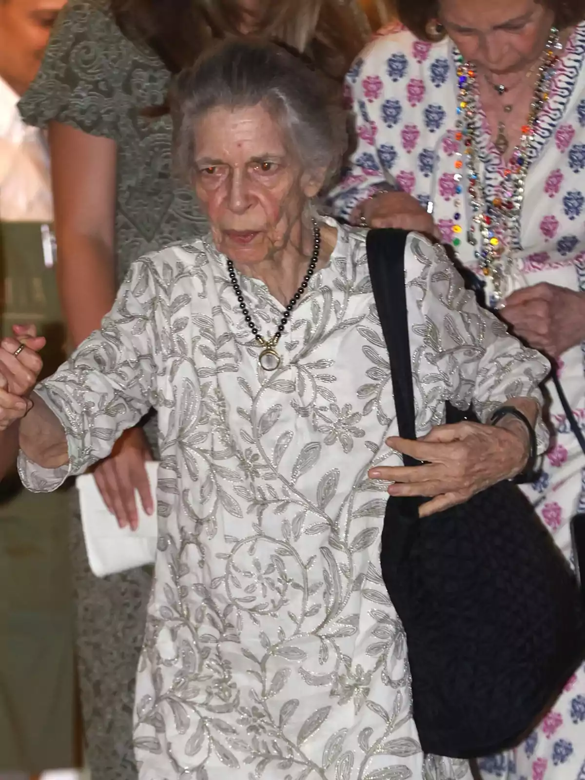 Mujer mayor de cabello canoso vestida con túnica blanca de bordados plateados y collar negro, caminando acompañada de otras personas Mujer mayor de cabello canoso vestida con túnica blanca de bordados plateados y collar negro, caminando acompañada de otras personas