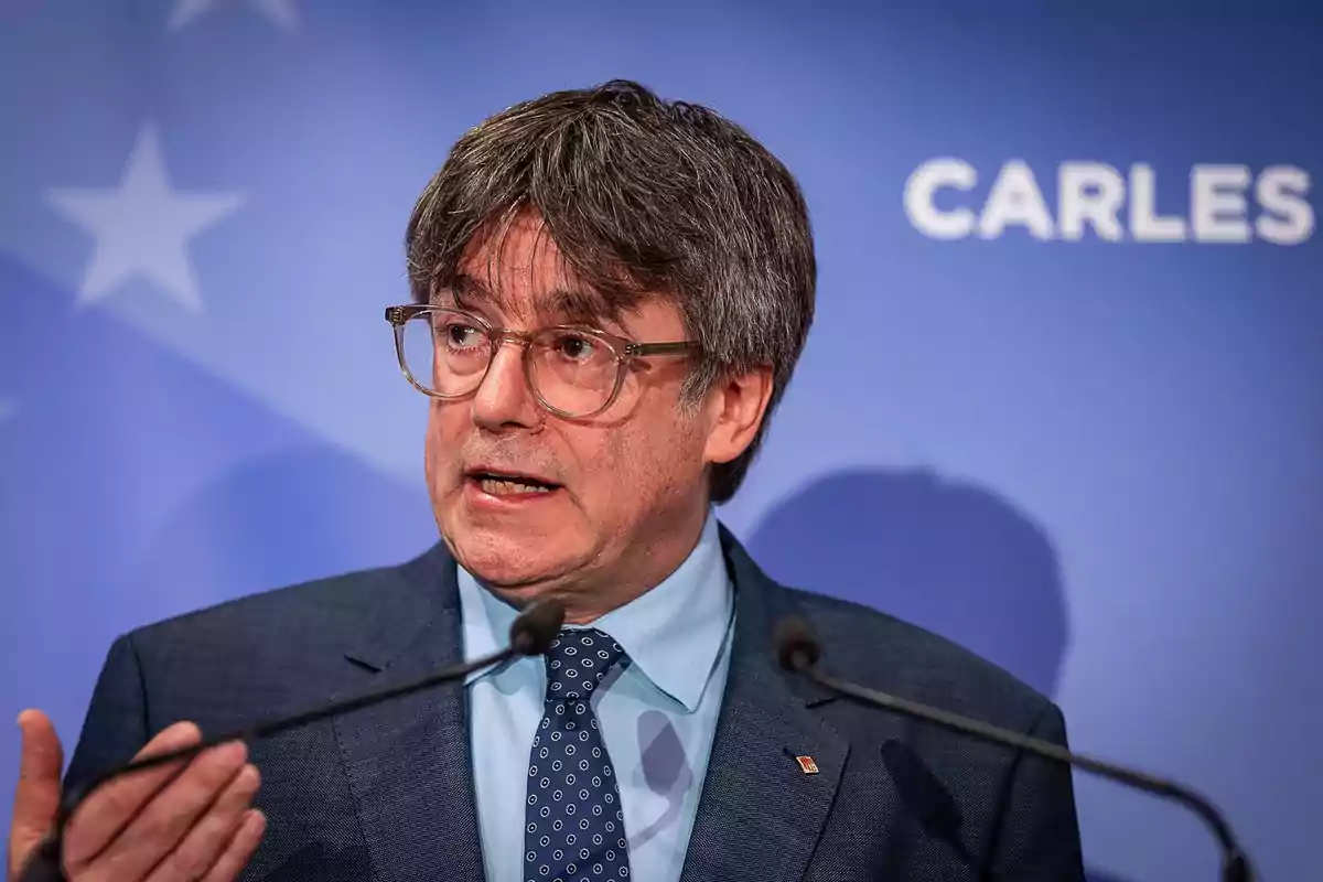 Hombre de cabello canoso y gafas hablando frente a micrófonos con fondo azul y letras blancas Hombre de cabello canoso y gafas hablando frente a micrófonos con fondo azul y letras blancas