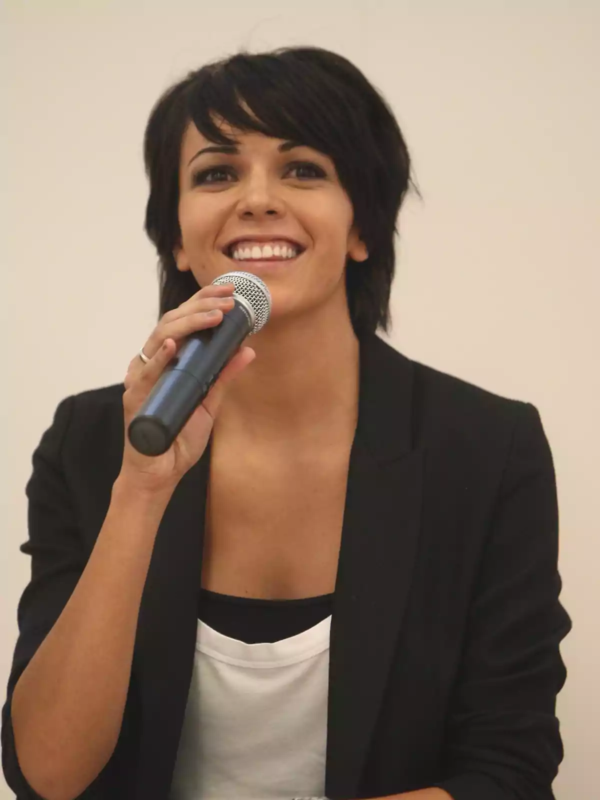 Mujer joven de cabello corto y oscuro sonriendo mientras sostiene un micrófono y viste un blazer negro sobre una camiseta blanca Mujer joven de cabello corto y oscuro sonriendo mientras sostiene un micrófono y viste un blazer negro sobre una camiseta blanca