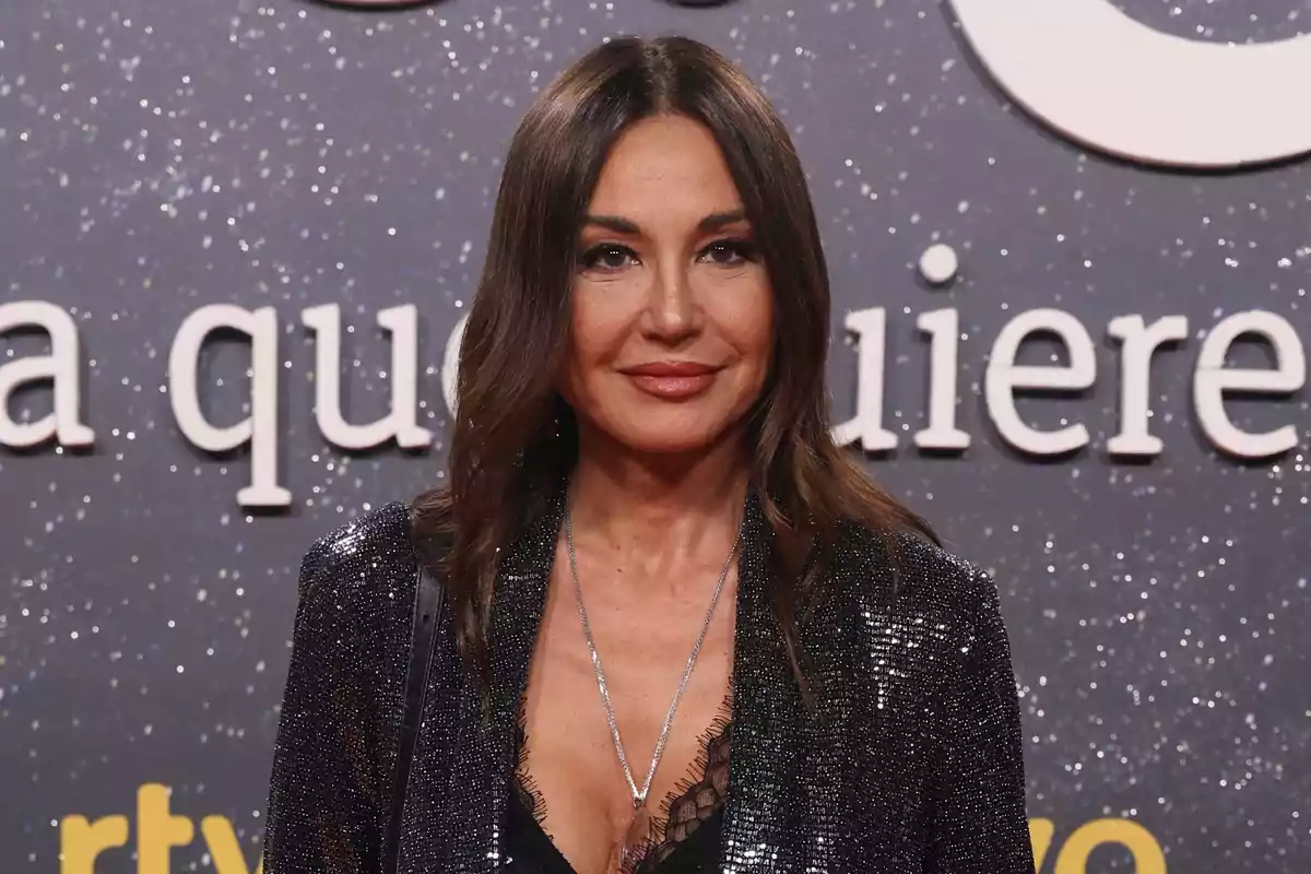 Mujer de cabello castaño y liso con chaqueta brillante posando frente a un fondo con letras grandes y destellos plateados