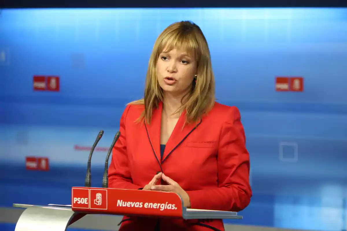 Mujer con chaqueta roja hablando en una rueda de prensa frente a un atril del PSOE con el lema Nuevas energías Mujer con chaqueta roja hablando en una rueda de prensa frente a un atril del PSOE con el lema Nuevas energías