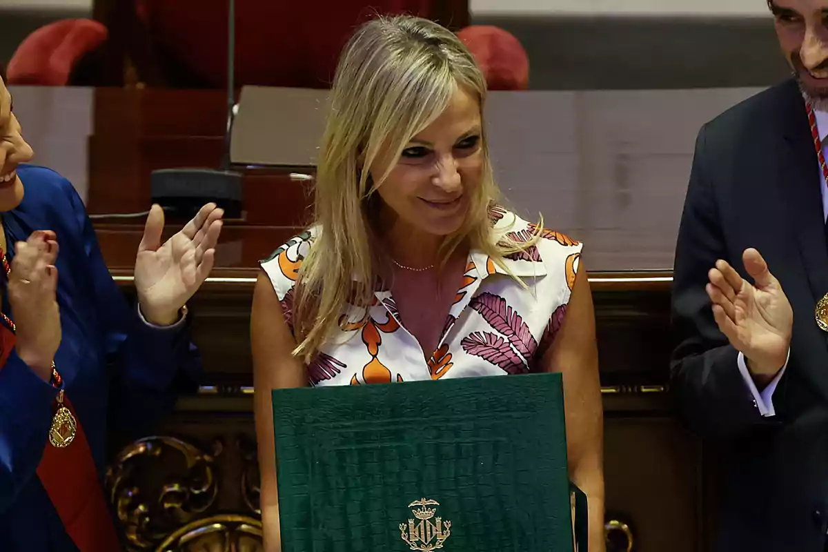 Mujer rubia sonriente con vestido estampado sostiene una carpeta verde mientras es aplaudida por personas a su alrededor en un entorno formal Mujer rubia sonriente con vestido estampado sostiene una carpeta verde mientras es aplaudida por personas a su alrededor en un entorno formal