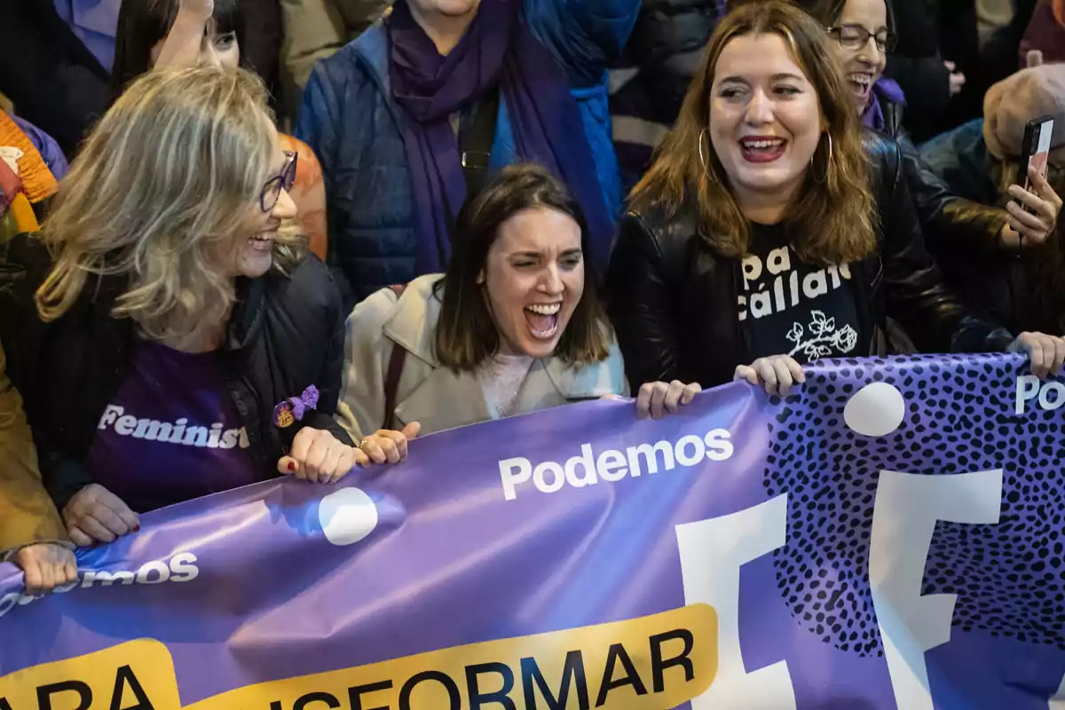 Tres mujeres sonríen y gritan con entusiasmo mientras sostienen una pancarta morada de Podemos en una manifestación feminista Tres mujeres sonríen y gritan con entusiasmo mientras sostienen una pancarta morada de Podemos en una manifestación feminista