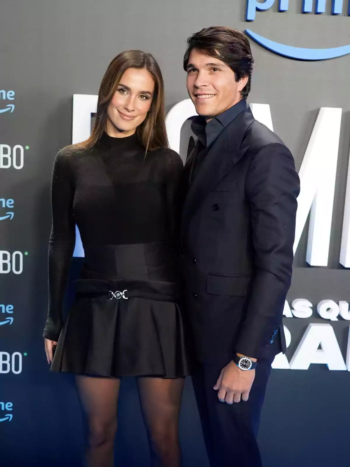 Una pareja posa sonriente en una alfombra azul durante un evento, ambos vestidos de negro y de manera elegante Una pareja posa sonriente en una alfombra azul durante un evento, ambos vestidos de negro y de manera elegante