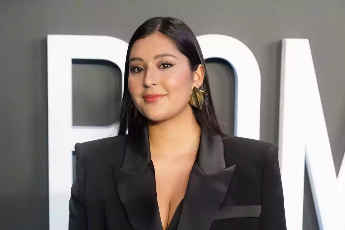 Mujer con cabello lacio y oscuro usando un saco negro elegante y aretes dorados grandes posando frente a un fondo gris con letras blancas grandes