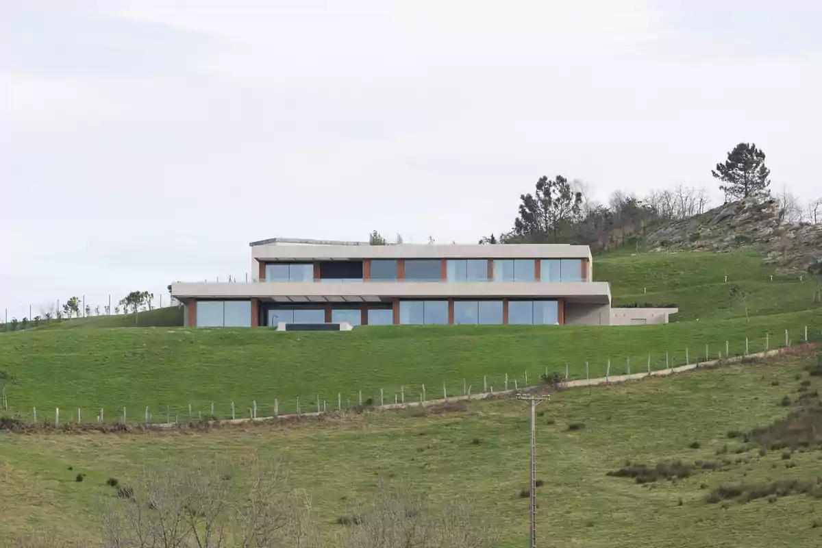 Casa moderna de dos plantas con grandes ventanales ubicada en una colina verde rodeada de naturaleza y árboles. Casa moderna de dos plantas con grandes ventanales ubicada en una colina verde rodeada de naturaleza y árboles.