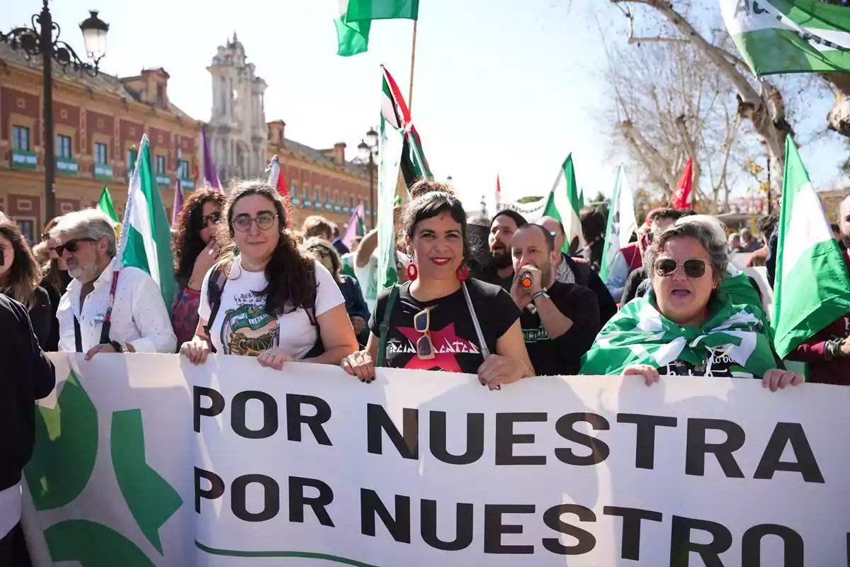 Personas manifestándose en la calle sosteniendo una pancarta y banderas verdes y blancas, con edificios históricos al fondo y ambiente soleado. Personas manifestándose en la calle sosteniendo una pancarta y banderas verdes y blancas, con edificios históricos al fondo y ambiente soleado.