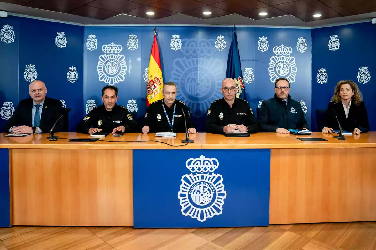 Seis personas sentadas detrás de una mesa en una sala de prensa de la Policía Nacional de España con banderas y logotipos oficiales al fondo