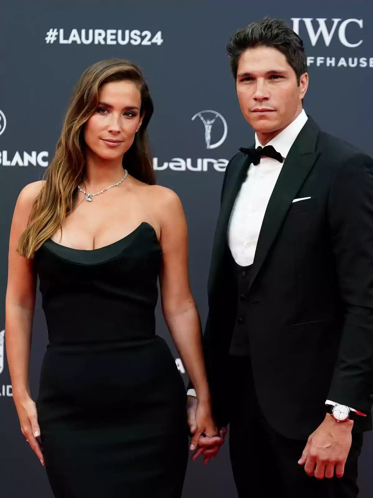 Una pareja elegantemente vestida posa tomada de la mano en una alfombra roja frente a un fondo con logotipos de un evento. Una pareja elegantemente vestida posa tomada de la mano en una alfombra roja frente a un fondo con logotipos de un evento.
