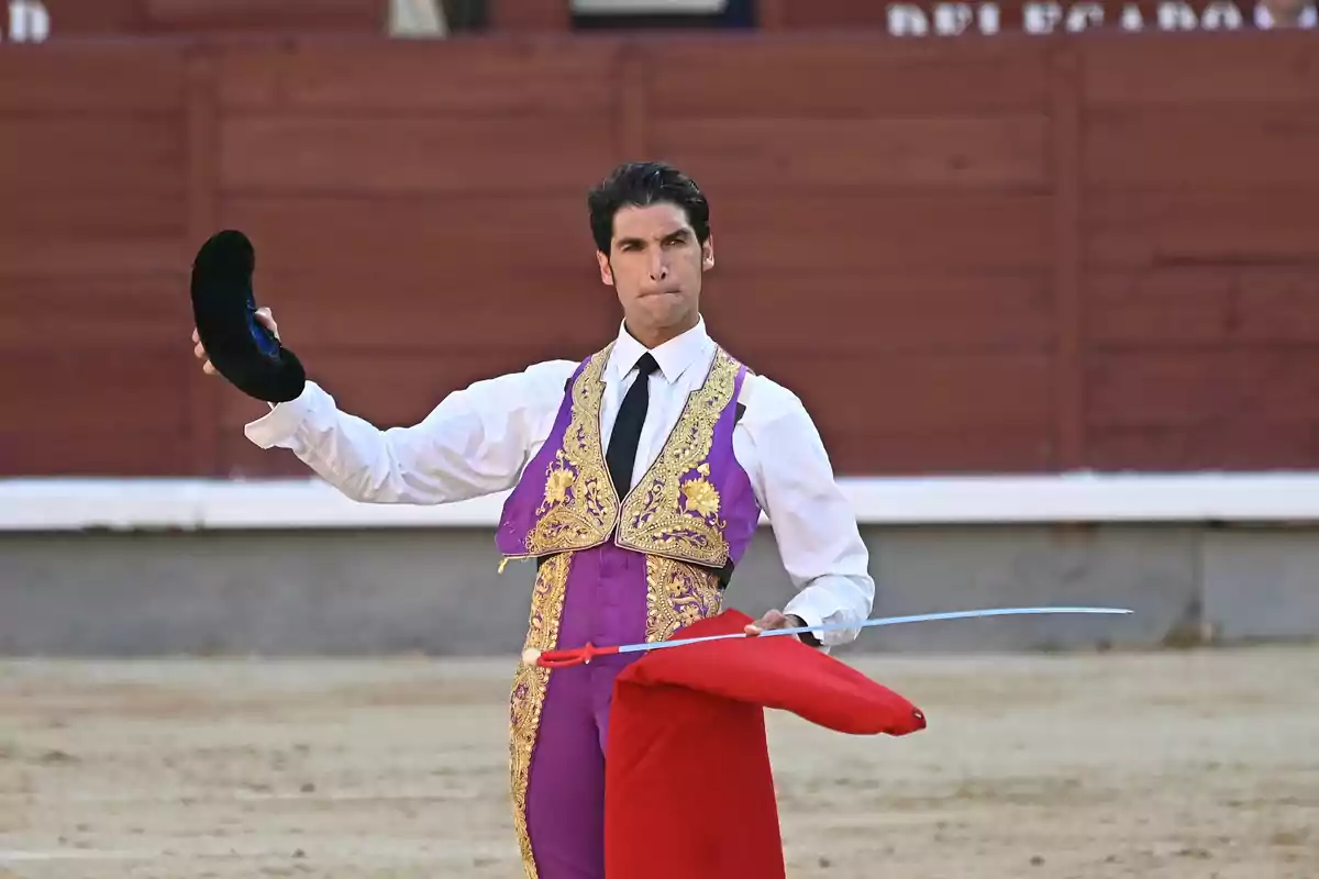 Torero en la plaza de toros sosteniendo la montera en una mano y la muleta y la espada en la otra, vestido con traje de luces morado y dorado