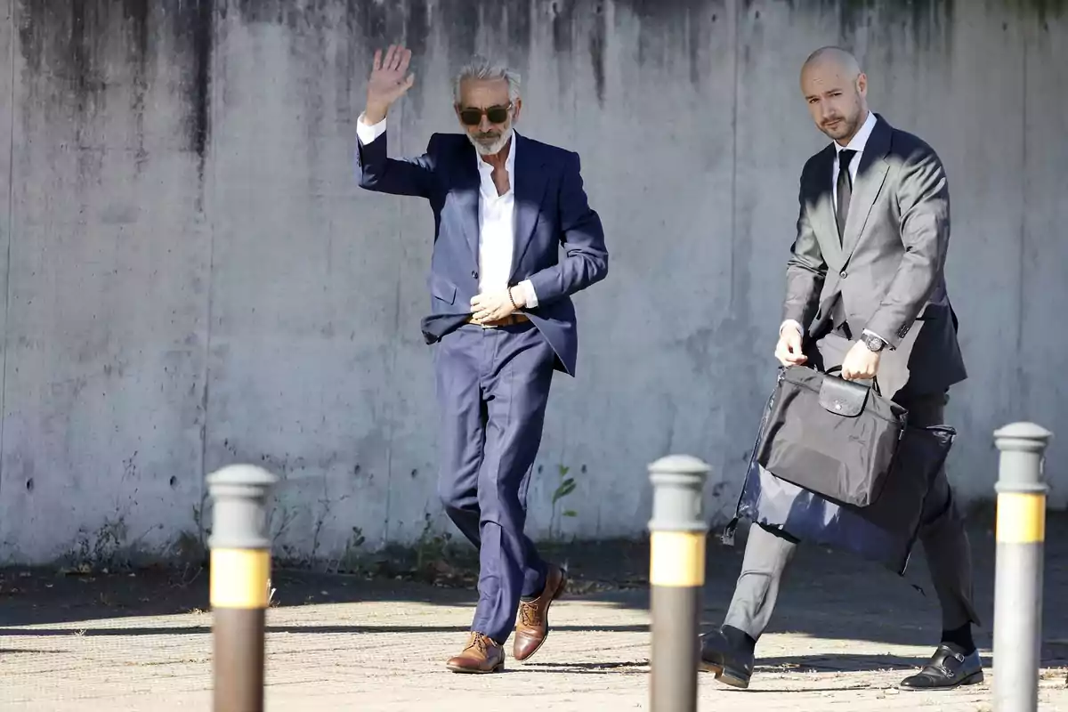 Dos hombres con traje caminan por la calle, uno de ellos saluda levantando la mano mientras el otro lleva un portafolio negro Dos hombres con traje caminan por la calle, uno de ellos saluda levantando la mano mientras el otro lleva un portafolio negro