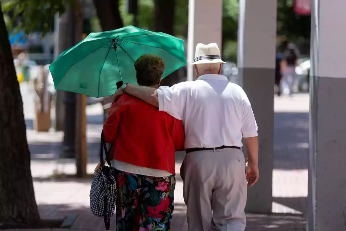 Pareja de personas mayores caminando juntas bajo una sombrilla verde en una acera Pareja de personas mayores caminando juntas bajo una sombrilla verde en una acera